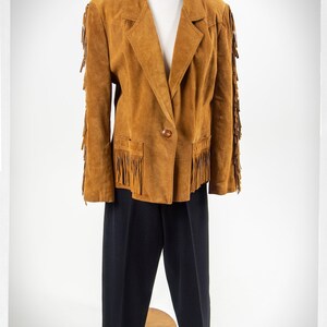 May include: A brown suede jacket with fringe detailing on the sleeves and pockets. The jacket has a single button closure and a classic blazer style.