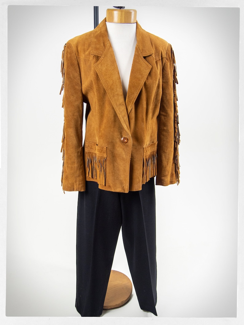 May include: A brown suede jacket with fringe detailing on the sleeves and pockets. The jacket has a single button closure and a classic blazer style.