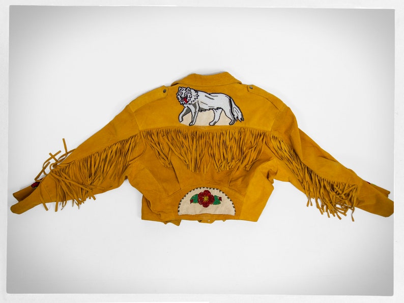 May include: A yellow suede jacket with fringe detailing and an embroidered wolf design on the back. The jacket also features a beaded flower design on the back.