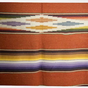May include: A woven blanket with a colorful striped pattern in shades of red, brown, yellow, purple, and white. The blanket has a diamond-shaped design in the center.