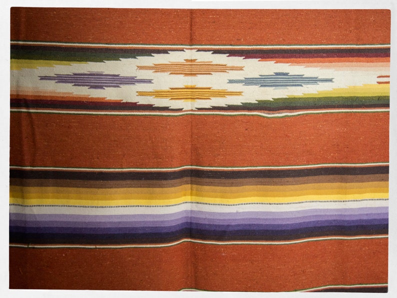 May include: A woven blanket with a colorful striped pattern in shades of red, brown, yellow, purple, and white. The blanket has a diamond-shaped design in the center.