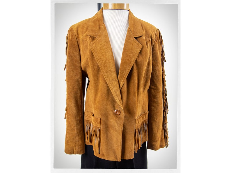 May include: A brown suede jacket with fringe detailing on the sleeves and hem. The jacket has a single button closure and a pocket on the left side.