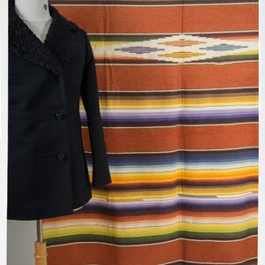 May include: A black peacoat with a brown fur collar is displayed on a mannequin in front of a colorful striped blanket with a geometric design in the center.