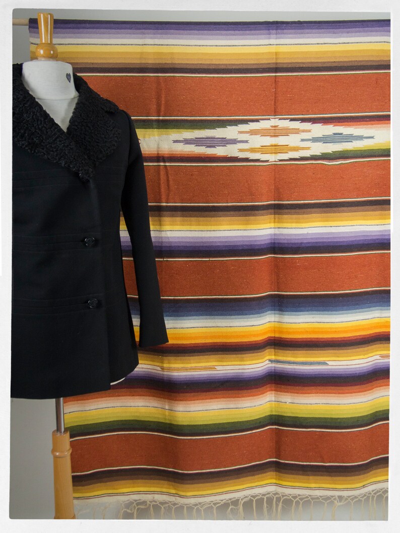 May include: A black peacoat with a brown fur collar is displayed on a mannequin in front of a colorful striped blanket with a geometric design in the center.