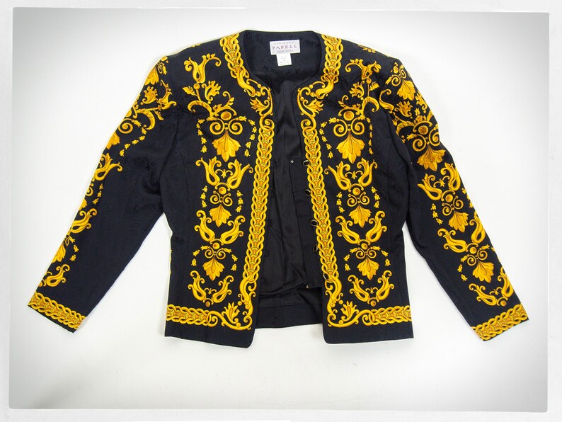Puede incluir: Una chaqueta negra con bordados dorados con un patr&oacute;n floral. La chaqueta tiene un cuello mandar&iacute;n y un cierre de un solo bot&oacute;n.