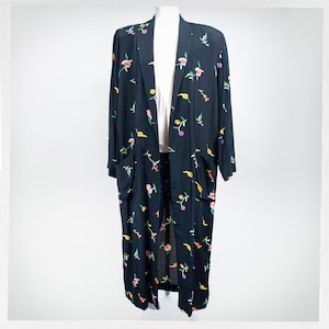 May include: A long, black open-front duster coat featuring a scattered floral pattern in shades of pink, yellow, and green. The garment includes long sleeves, a straight collarless neckline, and two large patch pockets on the front panels.