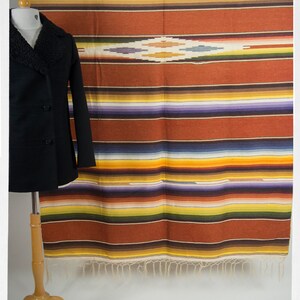 May include: A colorful striped blanket with a fringe border hangs on a wall. The blanket has a diamond pattern in the center. A black coat is displayed on a wooden stand in front of the blanket.