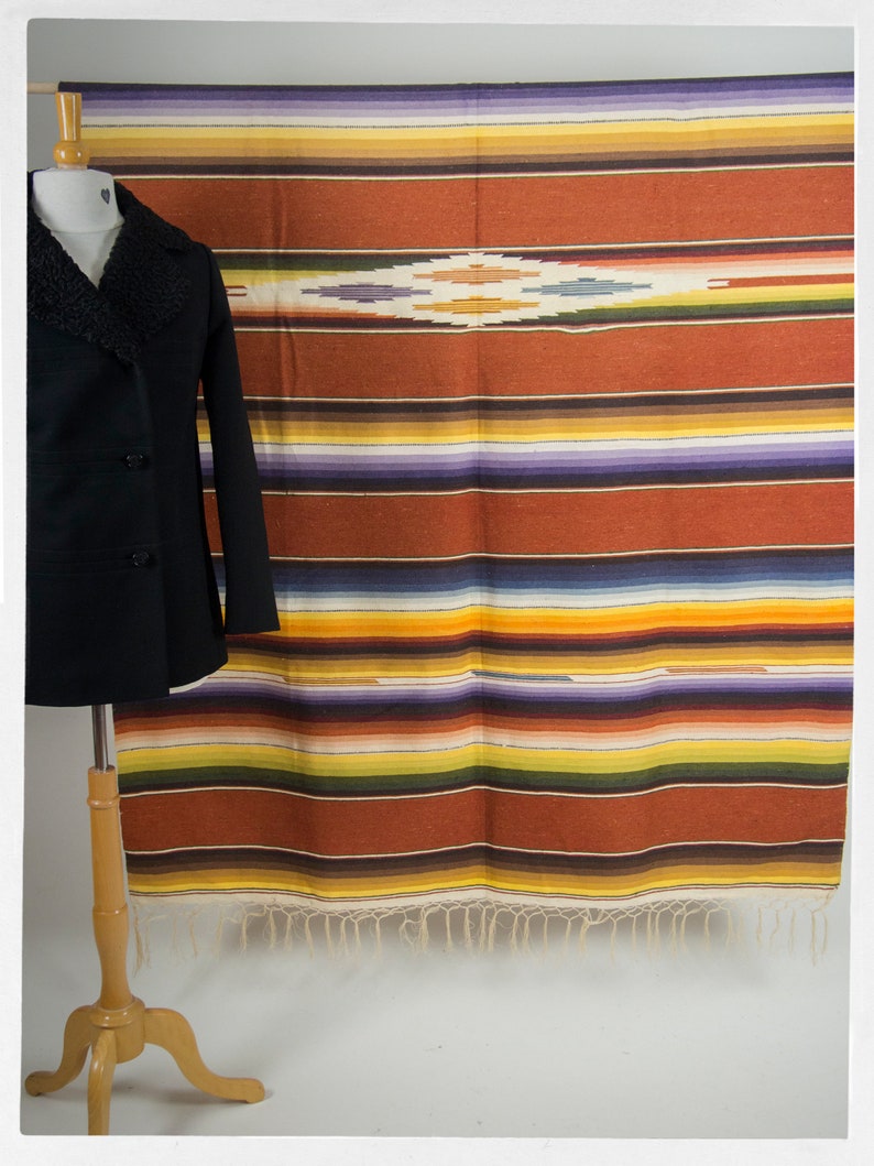 May include: A colorful striped blanket with a fringe border hangs on a wall. The blanket has a diamond pattern in the center. A black coat is displayed on a wooden stand in front of the blanket.