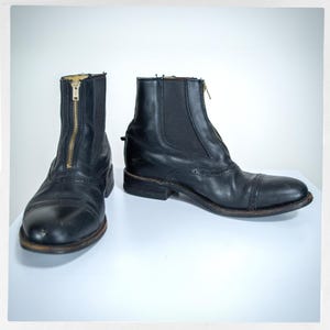 May include: Black leather ankle boots with a front zipper and elastic side panels. The boots have a cap toe design and a low heel. The zipper is gold-colored. The boots are displayed on a white surface.