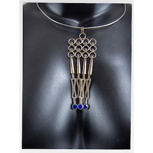 Puede incluir: Collar con colgante plateado con un dise&ntilde;o geom&eacute;trico. El colgante presenta una serie de c&iacute;rculos, barras verticales y formas que se cruzan, acentuadas con peque&ntilde;as piedras circulares azules. El collar se exhibe en un maniqu&iacute; negro.