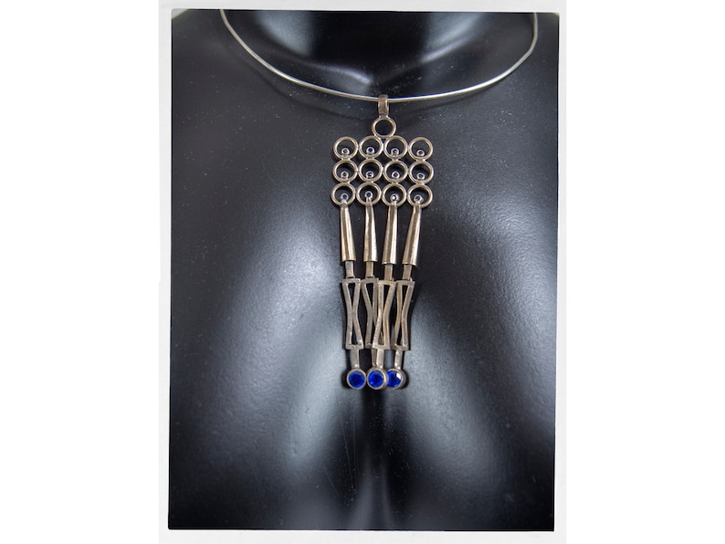 Puede incluir: Collar con colgante plateado con un dise&ntilde;o geom&eacute;trico. El colgante presenta una serie de c&iacute;rculos, barras verticales y formas que se cruzan, acentuadas con peque&ntilde;as piedras circulares azules. El collar se exhibe en un maniqu&iacute; negro.