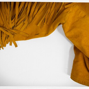 May include: A close-up of a mustard yellow suede jacket with fringe detail. The jacket is folded and the fringe is visible on the left side of the image.