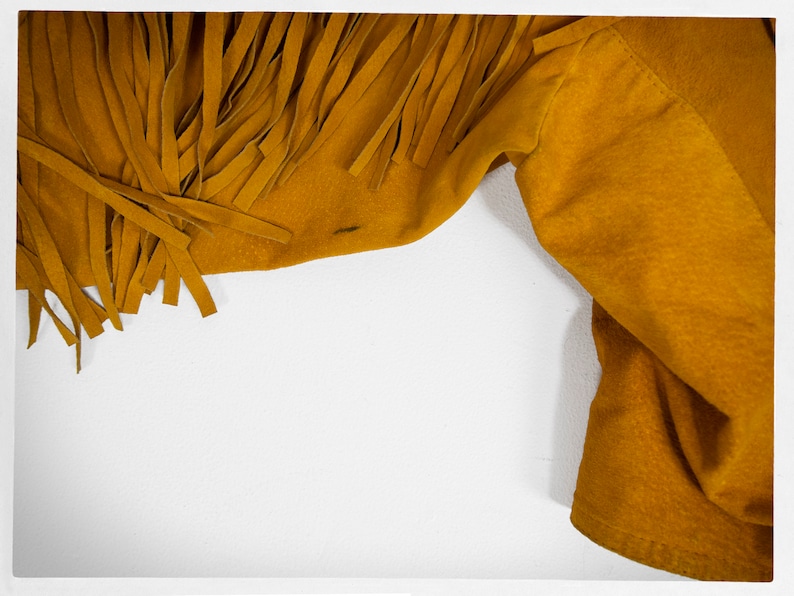 May include: A close-up of a mustard yellow suede jacket with fringe detail. The jacket is folded and the fringe is visible on the left side of the image.
