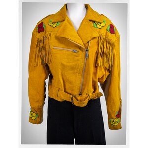 May include: A mustard yellow suede jacket with fringe detailing and embroidered floral accents. The jacket has a zipper closure and a belt.