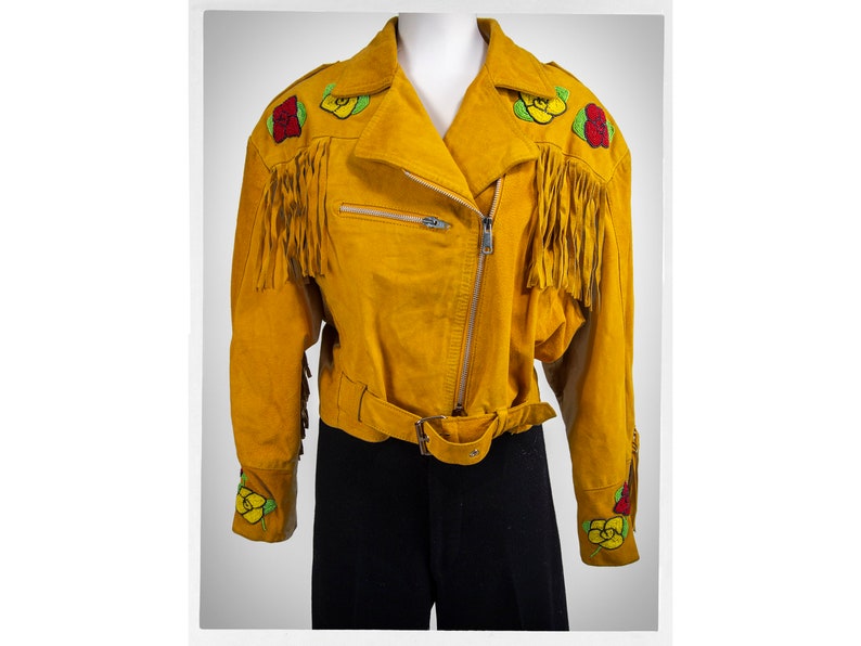 May include: A mustard yellow suede jacket with fringe detailing and embroidered floral accents. The jacket has a zipper closure and a belt.