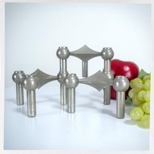 May include: A silver-toned metal candle holder with six candle cups. The design features geometric shapes and a mid-century modern aesthetic. A red apple and a bunch of green grapes are in the background.