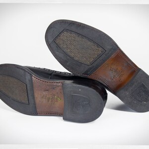 Op de afbeelding: Close-up van de zolen van een paar zwarte leren nette schoenen. De schoenen hebben een textuurrubberen zool en een bruine leren hak.