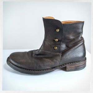 May include: A brown leather ankle boot with three decorative buttons on the side. The boot has a rounded toe and a low heel. The leather appears worn, giving it a vintage look. The sole is stitched with visible thread.