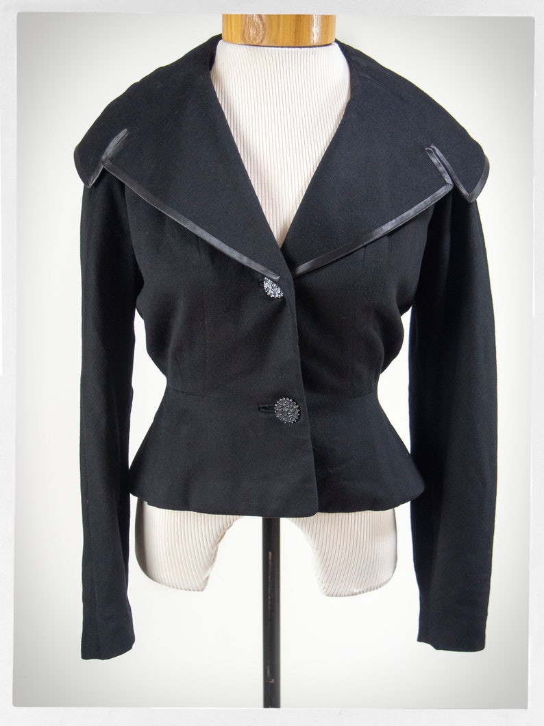 May include: A black cropped blazer with a wide, pointed collar and a fitted waist. The blazer has a satin trim and decorative buttons.
