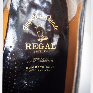 Op de afbeelding: Close-up van de binnenkant van een zwarte leren schoen met een gouden label met de tekst "Imperial Grade REGAL SINCE 1954 TRADITIONAL CLASSIC HANDCRAFTS KUM KANG SHOE MFG.CO., LTD."
