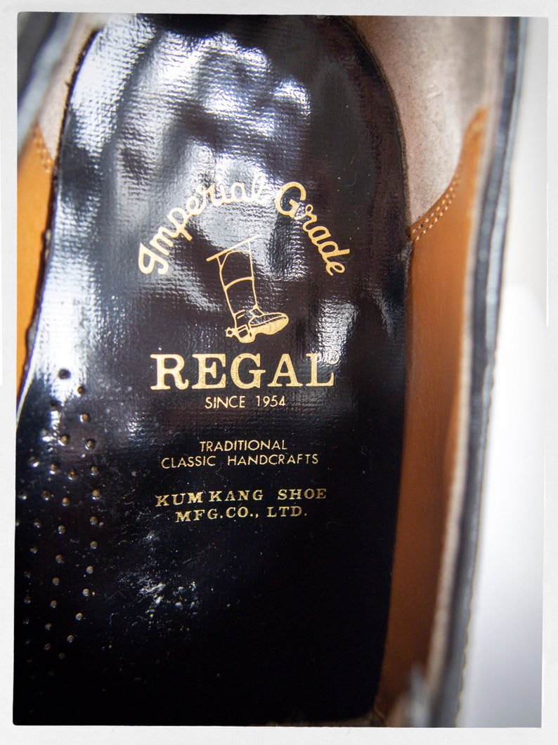 Op de afbeelding: Close-up van de binnenkant van een zwarte leren schoen met een gouden label met de tekst "Imperial Grade REGAL SINCE 1954 TRADITIONAL CLASSIC HANDCRAFTS KUM KANG SHOE MFG.CO., LTD."