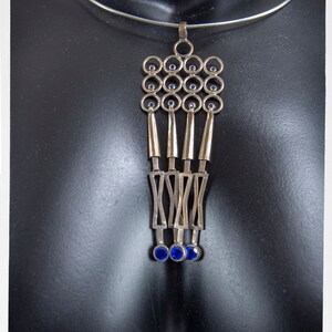 Puede incluir: Collar con colgante plateado con un dise&ntilde;o geom&eacute;trico. El colgante presenta una serie de c&iacute;rculos, formas alargadas y detalles de piedras preciosas azules. El collar se muestra en un maniqu&iacute; negro.