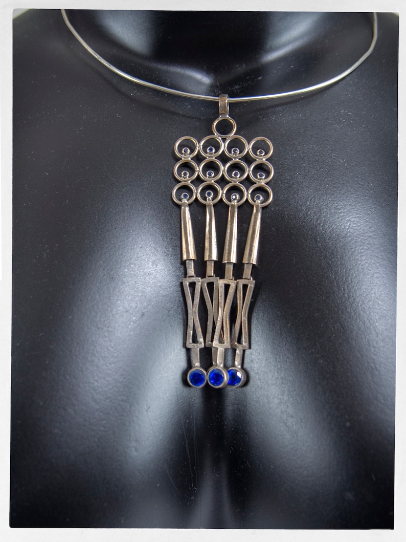 Puede incluir: Collar con colgante plateado con un dise&ntilde;o geom&eacute;trico. El colgante presenta una serie de c&iacute;rculos, formas alargadas y detalles de piedras preciosas azules. El collar se muestra en un maniqu&iacute; negro.