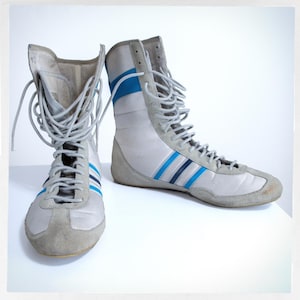 May include: A pair of white high-top boxing shoes with blue and black stripes. The shoes have laces and are made of leather.