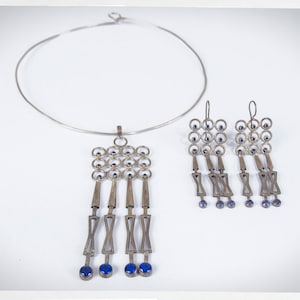 Puede incluir: Un conjunto de collar y pendientes plateados. El collar es de estilo gargantilla. El colgante y los pendientes presentan un dise&ntilde;o geom&eacute;trico con peque&ntilde;as piedras azules y elementos colgantes. Las joyas se exhiben sobre un fondo blanco.
