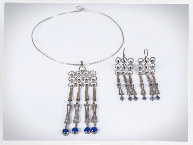 Puede incluir: Un conjunto de collar y pendientes plateados. El collar es de estilo gargantilla. El colgante y los pendientes presentan un dise&ntilde;o geom&eacute;trico con peque&ntilde;as piedras azules y elementos colgantes. Las joyas se exhiben sobre un fondo blanco.
