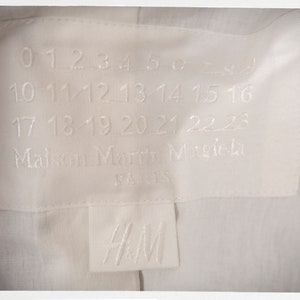 May include: White clothing label with black text that reads "0 1 2 3 4 5 6 7 8 9 10 11 12 13 14 15 16 17 18 19 20 21 22 23 Maison Margiela PARIS" and a white label with black text that reads "H&M".