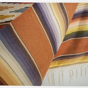 May include: A woven rug with a striped pattern in shades of brown, yellow, white, and purple. The rug has a fringe on the bottom edge.
