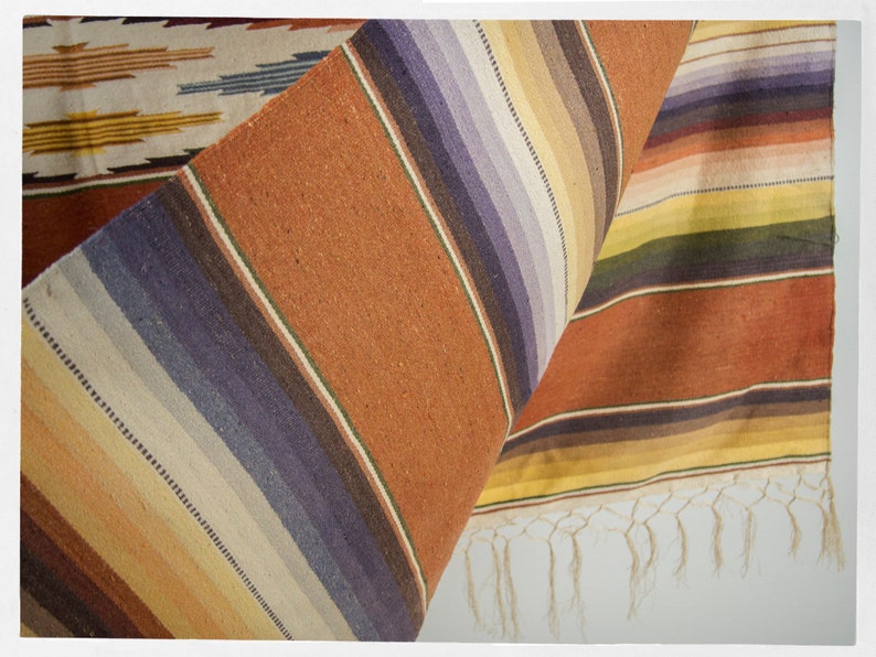 May include: A woven rug with a striped pattern in shades of brown, yellow, white, and purple. The rug has a fringe on the bottom edge.