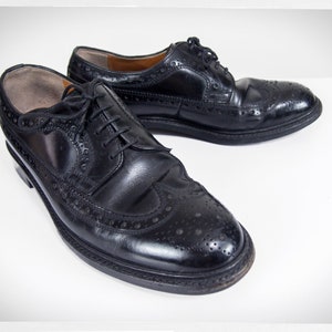 Op de afbeelding: Een paar zwarte leren oxfords met brogue details. De schoenen hebben een vetersluiting en een ronde neus.