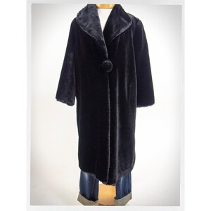 May include: A long, black faux fur coat with a large collar and a single button closure. The coat is displayed on a mannequin, showcasing its full length and design. The coat's texture appears soft and plush.