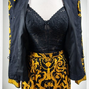 Puede incluir: Un traje de falda negro y dorado con estampado, con un top de encaje negro debajo. La chaqueta tiene un estampado paisley dorado y la falda tiene un estampado floral dorado.
