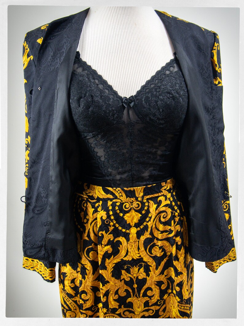 Puede incluir: Un traje de falda negro y dorado con estampado, con un top de encaje negro debajo. La chaqueta tiene un estampado paisley dorado y la falda tiene un estampado floral dorado.