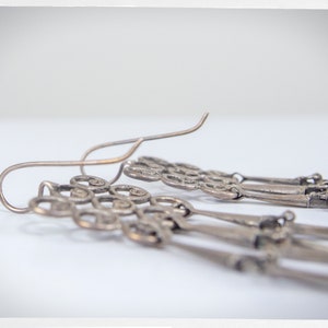 Puede incluir: Primer plano de pendientes colgantes plateados. Los pendientes presentan un dise&ntilde;o decorativo con m&uacute;ltiples hilos de abalorios ornamentados en forma de llave. Los ganchos para las orejas son curvos y sencillos.