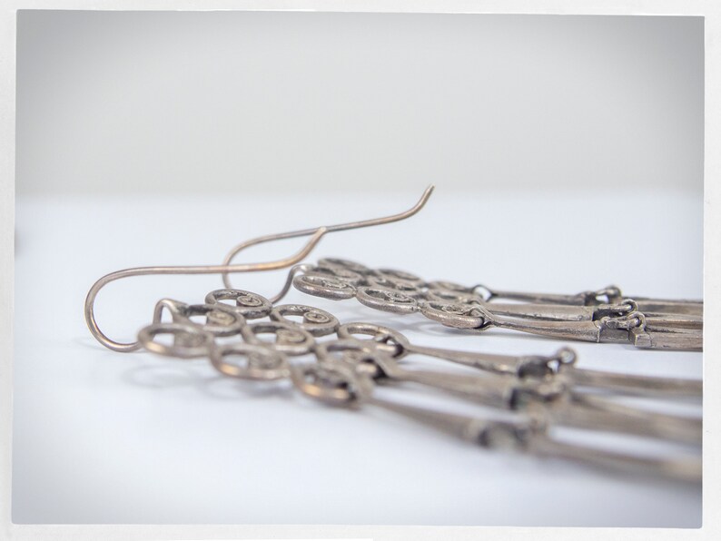 Puede incluir: Primer plano de pendientes colgantes plateados. Los pendientes presentan un dise&ntilde;o decorativo con m&uacute;ltiples hilos de abalorios ornamentados en forma de llave. Los ganchos para las orejas son curvos y sencillos.