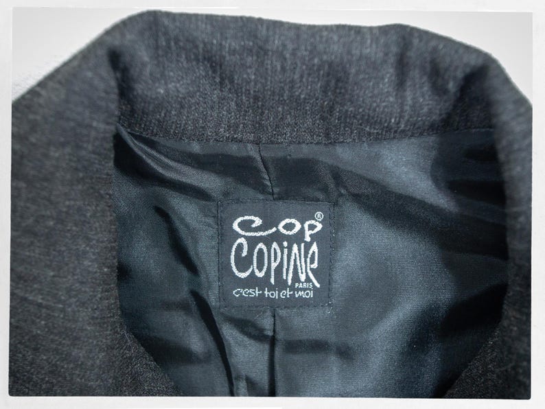 May include: Close-up of a dark gray blazer collar with a black satin lining. The lining features a white label with the brand name "Copine Paris" and the phrase "c'est toi et moi".