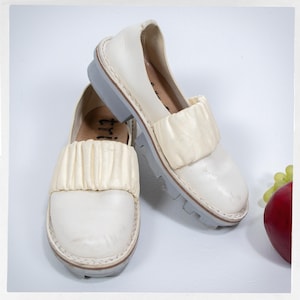 May include: Off-white leather slip-on shoes with a gathered fabric detail across the top. The shoes have a gray sole and a rounded toe. The image shows two shoes, one slightly in front of the other, on a white surface.