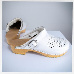 May include: White leather clogs with a wooden sole and a buckle closure. The clogs have a perforated design on the top and a light brown wooden base. The sole is a light yellow color. One clog is in the background, and the other is in the foreground.