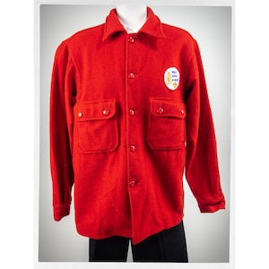 May include: A red wool shirt with a patch that says "Bull Dog Scout Achiever" on the left chest. The shirt has two large pockets on the front and a button-up closure.
