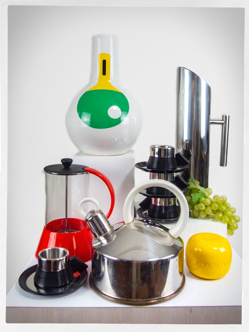 K&ouml;nnte beinhalten: Ein Set K&uuml;chenutensilien mit einer wei&szlig;en Keramikvase mit gr&uuml;nem und gelbem Design, einer silbernen Kanne, einer roten und klaren Glas-French-Press, einem silbernen Wasserkocher, einer wei&szlig;en Keramiktasse und Untertasse und einem gelben Dekorationsobjekt.