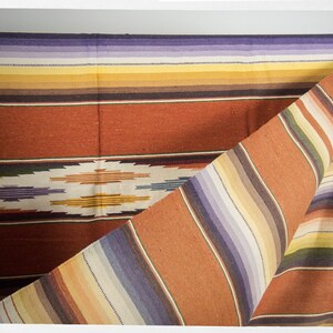May include: A woven blanket with a colorful striped pattern in shades of orange, yellow, purple, and brown. The blanket is folded in half, revealing the intricate design.