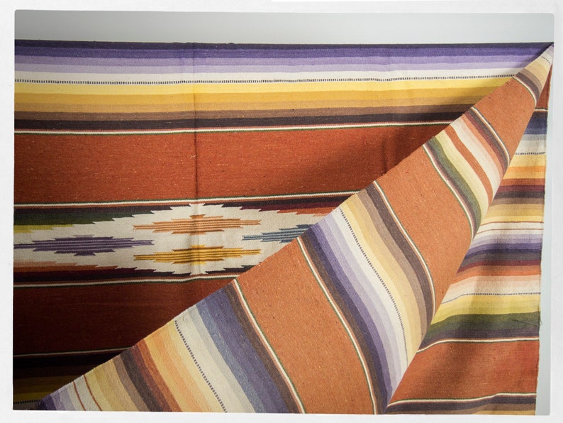 May include: A woven blanket with a colorful striped pattern in shades of orange, yellow, purple, and brown. The blanket is folded in half, revealing the intricate design.