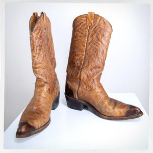 May include: A pair of vintage cowboy boots. The boots are made of distressed brown leather with a textured pattern. They have pointed toes and dark brown accents on the heels and toes. The boots are displayed on a white surface.