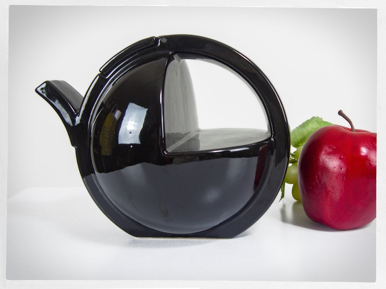 K&ouml;nnte beinhalten: Eine schwarze Teekanne aus Keramik mit einem einzigartigen Design. Die Teekanne hat einen runden Griff und einen Ausguss, der sich nach oben kr&uuml;mmt. Die Teekanne steht auf einer wei&szlig;en Oberfl&auml;che.