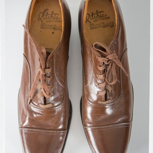 May include: A pair of brown leather lace-up shoes with a cap toe. The shoes have a label inside that reads "Ritchie Shoes for Men Master Built".