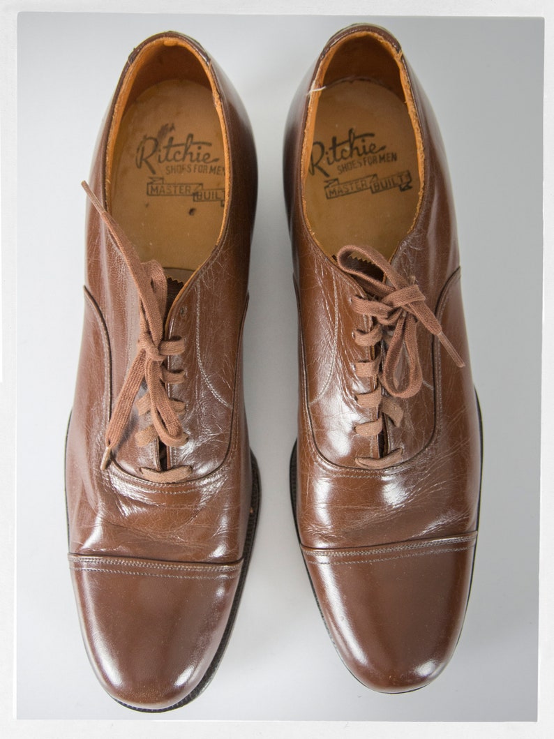 May include: A pair of brown leather lace-up shoes with a cap toe. The shoes have a label inside that reads "Ritchie Shoes for Men Master Built".